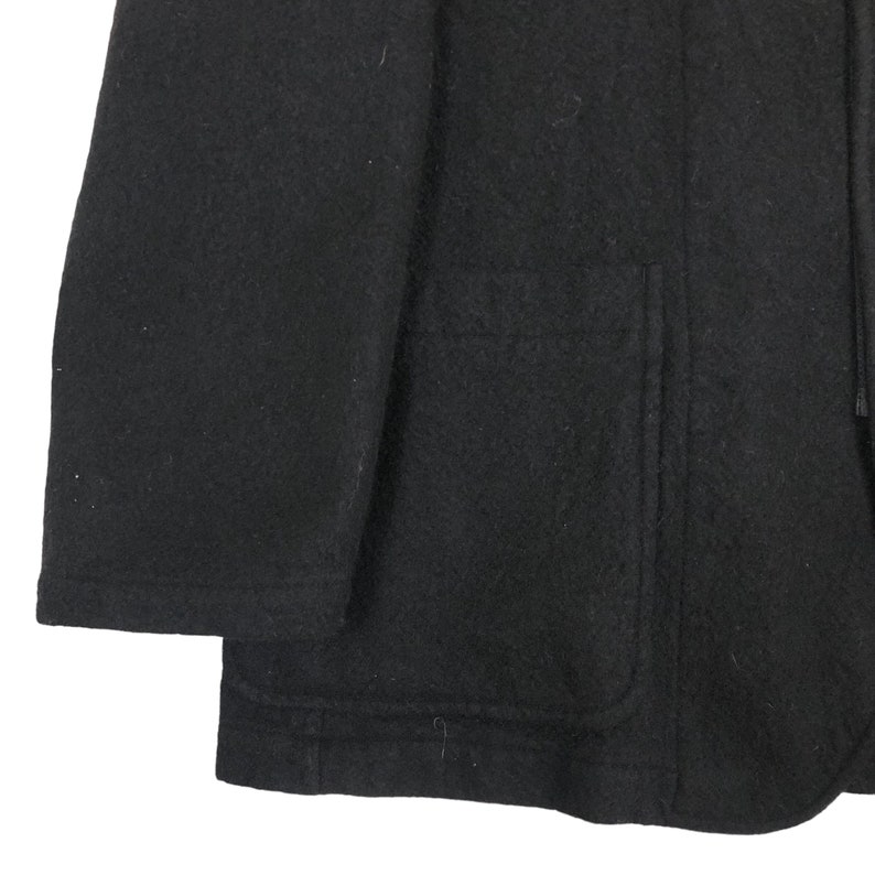 May include: A close-up of a black wool coat. The coat has a large pocket and a hood. The fabric appears to be thick and warm, suitable for cold weather. The coat is a solid black color.