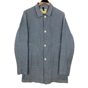 Puede incluir: Chaqueta a rayas azules y blancas con cuello y cierre de botones. La chaqueta tiene dos bolsillos delanteros y una etiqueta en el cuello. La chaqueta está colgada de una percha de madera.