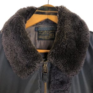 May include: Close-up of a vintage military-style jacket. The jacket features a dark gray exterior with a brown faux fur collar. A metal zipper runs down the front. The jacket is hanging on a wooden hanger.