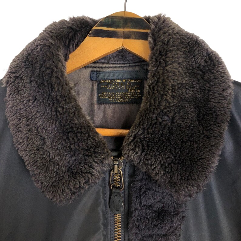 May include: Close-up of a vintage military-style jacket. The jacket features a dark gray exterior with a brown faux fur collar. A metal zipper runs down the front. The jacket is hanging on a wooden hanger.