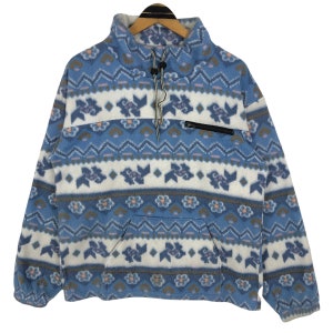 Op de afbeelding: Een blauwe, witte en patroon fleece trui met een hoge kraag en een klein zakje met rits. Het ontwerp bevat bloemen en geometrische vormen in blauw, roze en bruin. De trui heeft een trekkoord sluiting bij de nek. Afmetingen in centimeters.