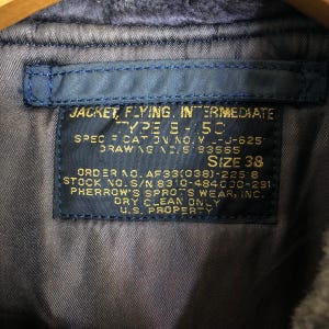 May include: Close-up of a blue label sewn into a jacket. The label reads "JACKET, FLYING, INTERMEDIATE TYPE B-5C" and includes size 38. Additional text includes "U.S. PROPERTY" and other specifications.