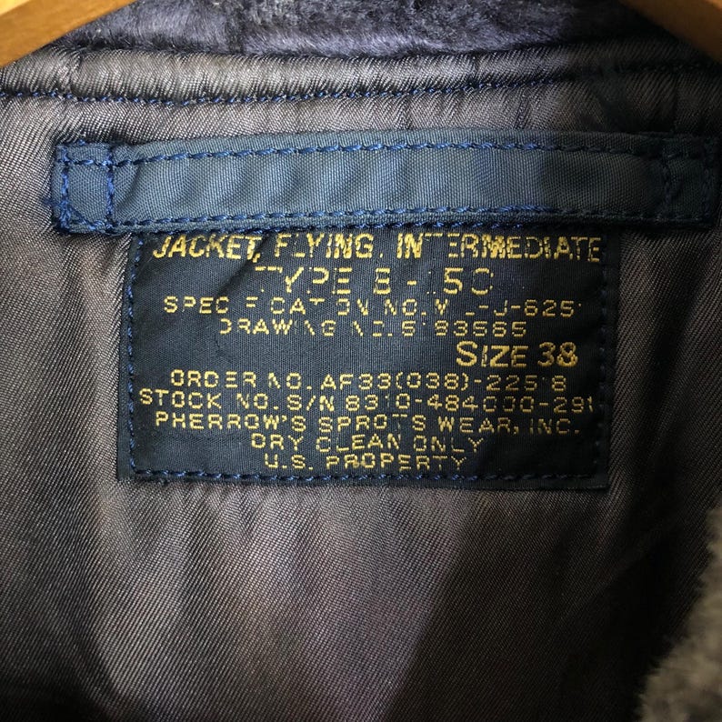 May include: Close-up of a blue label sewn into a jacket. The label reads "JACKET, FLYING, INTERMEDIATE TYPE B-5C" and includes size 38. Additional text includes "U.S. PROPERTY" and other specifications.