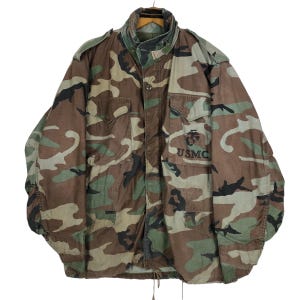 Puede incluir: Chaqueta militar de camuflaje con el texto "USMC" en el bolsillo delantero. La chaqueta tiene cierre de botones, múltiples bolsillos y cuello alto. El patrón incluye tonos de marrón, verde y beige.