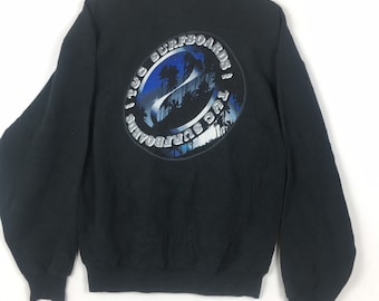 May include: Black sweatshirt with a circular graphic on the back. The graphic features a blue and white design with palm trees and the text "1 TUG SURFBOARDS". The sweatshirt has long sleeves and a crew neck.