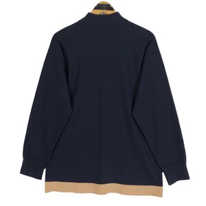 May include: Navy blue long-sleeve sweater with a mock neck and a tan bottom hem. The sweater is hanging on a wooden hanger against a white background. The sweater's design is simple and classic.