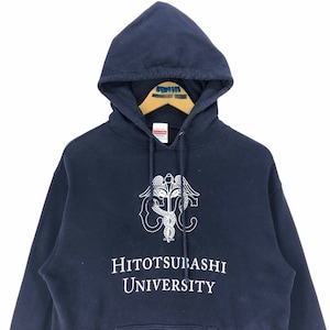 May include: A navy blue hoodie with a white emblem and text. The emblem is a caduceus design with wings and a serpent. The text reads "HITOTSUBASHI UNIVERSITY". The sweatshirt has a drawstring hood and is made of a thick material.