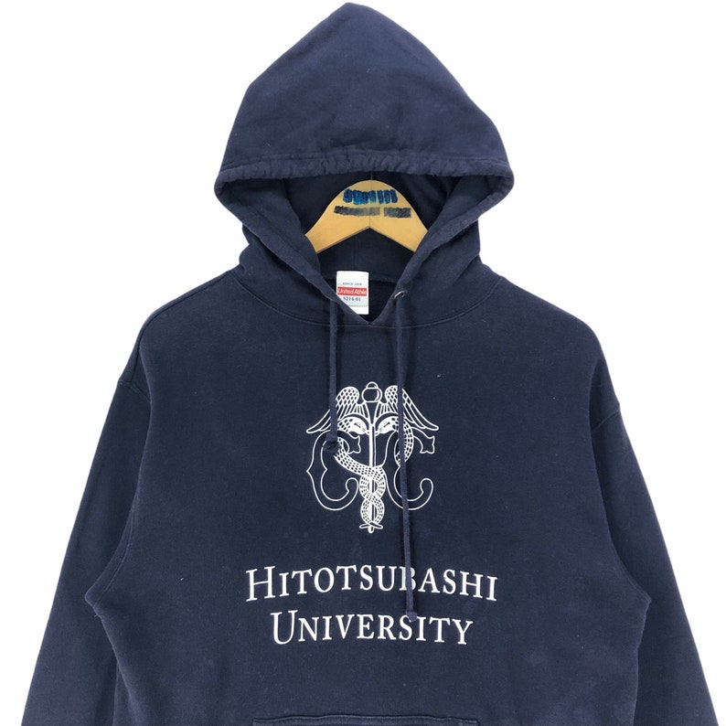 May include: A navy blue hoodie with a white emblem and text. The emblem is a caduceus design with wings and a serpent. The text reads "HITOTSUBASHI UNIVERSITY". The sweatshirt has a drawstring hood and is made of a thick material.