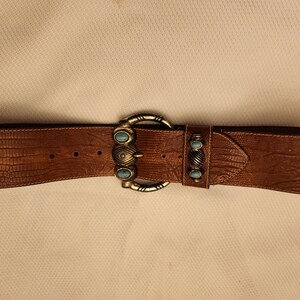 May include: Brown leather belt with a textured finish. The belt features a large silver-tone buckle with turquoise-colored accents. A smaller matching accent is on the belt loop. The belt is a wide style.