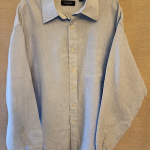 May include: A light blue button-down shirt with a collar and long sleeves. The shirt has a chest pocket and is hanging on a hanger. The fabric appears to be linen or a similar lightweight material. The shirt is unbuttoned.