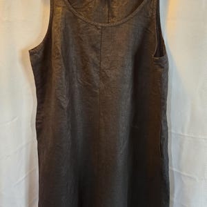May include: A dark charcoal-colored sleeveless dress with a scoop neckline and a relaxed fit. The dress has a simple, minimalist design with a vertical seam down the front and side pockets. It hangs on a black hanger against a white background.