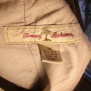 May include: A beige Tommy Bahama label with a palm tree logo and the text "Tommy Bahama". The label is attached to a garment with a tag that reads "85% Silk 15% Wool Made in China".