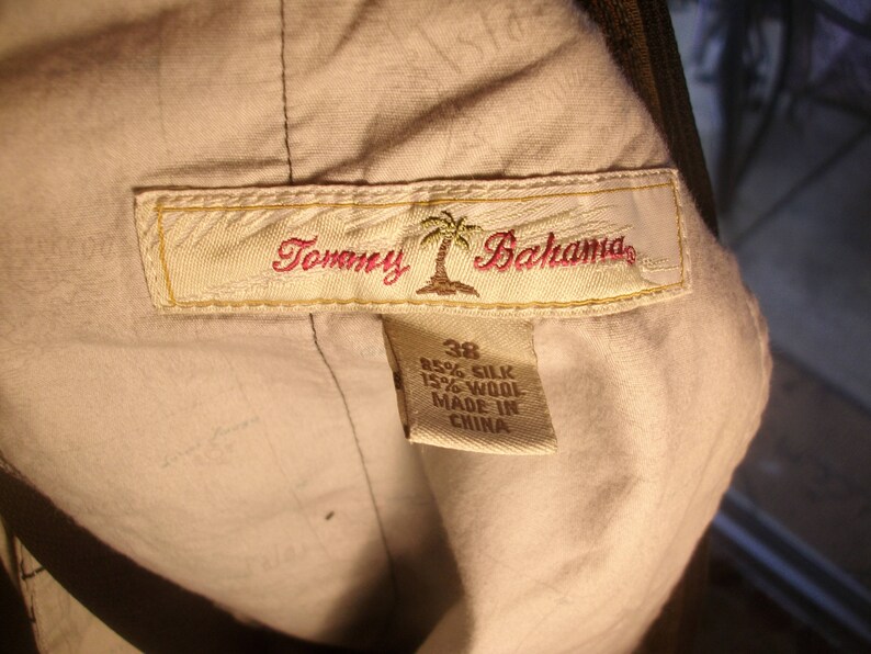 May include: A beige Tommy Bahama label with a palm tree logo and the text "Tommy Bahama". The label is attached to a garment with a tag that reads "85% Silk 15% Wool Made in China".