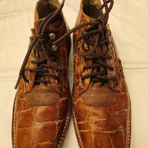 May include: A pair of brown leather boots with a crocodile skin pattern. The boots have dark brown laces, metal eyelets, and a square toe. The stitching is visible around the sole.