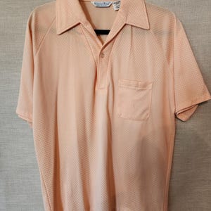 May include: Peach-coloured polo shirt with a textured dot pattern, short sleeves, and a chest pocket. The shirt has a classic collar and a three-button placket. The brand name "Carino Ford" is visible on the label.