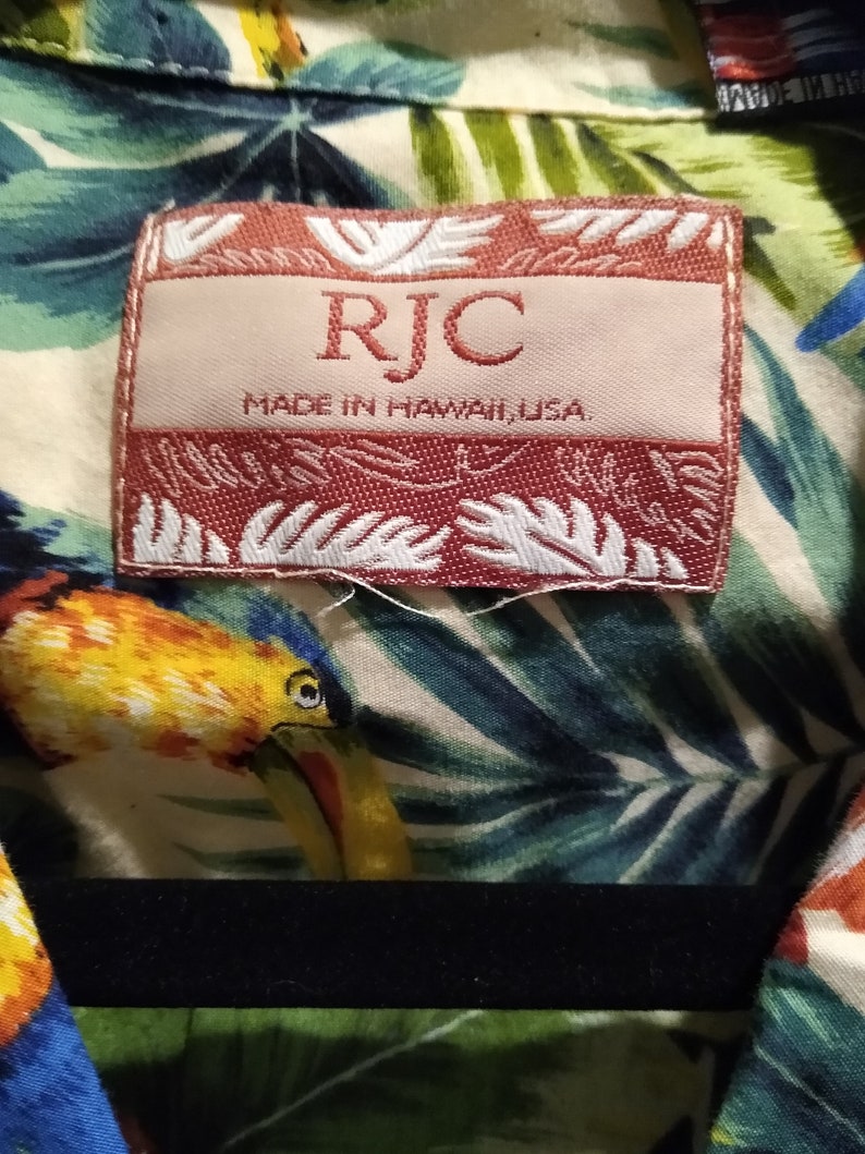 May include: A red and white label with the text "RJC" and "Made in Hawaii, USA". The label is attached to a white shirt with a tropical print.