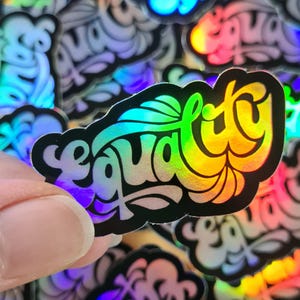 Könnte beinhalten: Ein holografischer Aufkleber mit dem Wort "Gleichheit" in einer stilisierten Schriftart. Das Wort ist mit einem Regenbogen-Farbverlauf gefüllt, und der Aufkleber hat einen schwarzen Umriss. Weitere ähnliche Aufkleber befinden sich im Hintergrund.