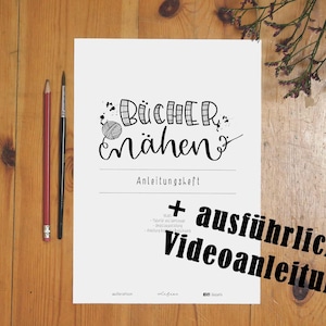 Op de afbeelding: Een wit vel papier met zwarte tekst die "Bücher nähen" en "Anleitungsheft + ausführlicher Videoanleitung" luidt. Het papier ligt op een houten oppervlak met een rode potlood en een zwart penseel.