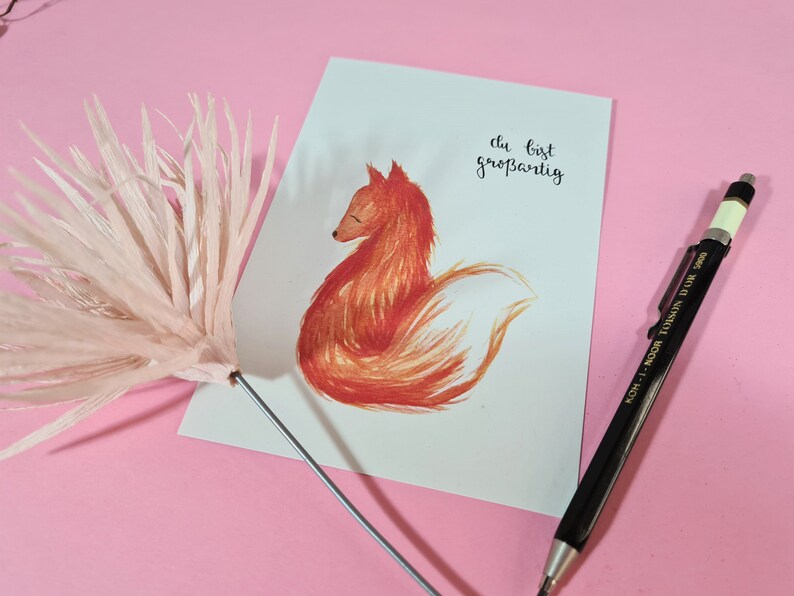 May include: A white card with a hand-drawn illustration of a red fox sitting with its tail curled around its body. The text "du bist gro&szlig;artig" is written in black ink on the card.