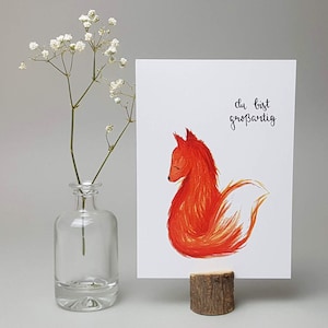 May include: A white card with a red fox illustration and the text "du bist gro&szlig;artig" in black calligraphy. The card is propped up on a small wooden stand.