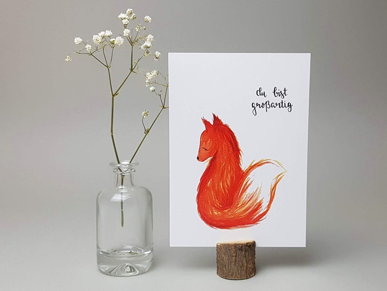 May include: A white card with a red fox illustration and the text "du bist gro&szlig;artig" in black calligraphy. The card is propped up on a small wooden stand.
