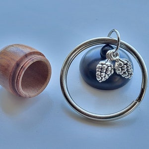 May include: A wooden acorn-shaped container with a removable lid sits next to a keychain. The keychain features a silver-colored ring, a black bead, and a charm with two silver-colored pine cones. The container is made of light brown wood.