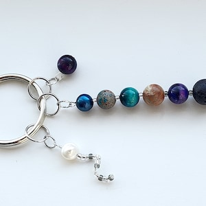 May include: A silver key chain with a variety of colorful beads, including blue, green, brown, purple, and black. The beads are attached to a silver ring and a silver chain with a white pearl.