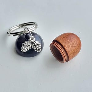 Puede incluir: Una pequeña caja de madera con forma de bellota con una tapa que se abre. La caja está unida a un llavero plateado con un dije plateado que parece una bellota.