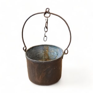 May include: A rustic, antique metal bucket with a rounded body and a curved handle. The bucket is suspended by a chain and has a weathered, aged appearance with shades of brown and blue-green. The handle is made of a thick, curved metal rod.