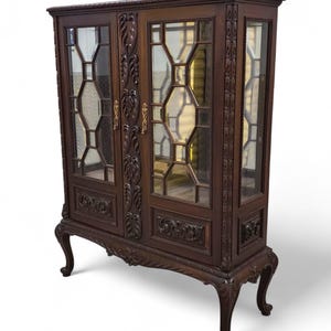 May include: A dark brown antique display cabinet with glass doors and ornate carvings. The cabinet features a rectangular shape, decorative legs, and gold-colored hardware. The glass panels have a geometric design.
