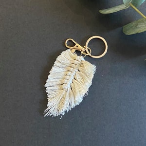 Puede incluir: Un llavero de macramé color crema con forma de pluma, que presenta un diseño central de cordón anudado con bordes deshilachados. La pieza textil está unida a un mosquetón de metal dorado y a una anilla circular, formando un accesorio.