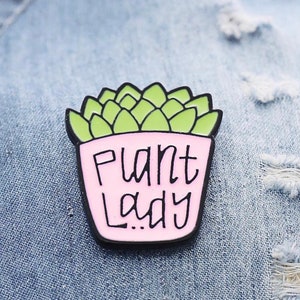 Puede incluir: Un pin de esmalte met&aacute;lico con forma de maceta rosa claro que contiene una planta suculenta de color verde claro. La maceta presenta el texto negro "PLANT LADY" escrito en un estilo casual y manual. El dise&ntilde;o est&aacute; delineado en metal negro.