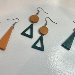 Puede incluir: Tres pares de pendientes colgantes con ganchos de metal plateado. Un par presenta triángulos de color naranja sólido, un par triángulos de color verde oscuro y un par combina un pequeño círculo naranja sobre un triángulo verde oscuro.