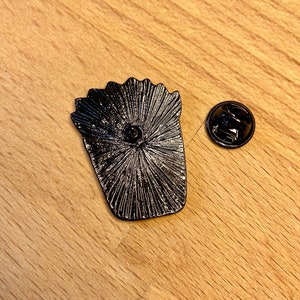 Puede incluir: Un pin de metal negro con forma de recipiente de patatas fritas, que muestra el reverso texturizado con un poste central y una pieza de cierre circular de metal negro separada, ambos con un acabado oscuro y mate.