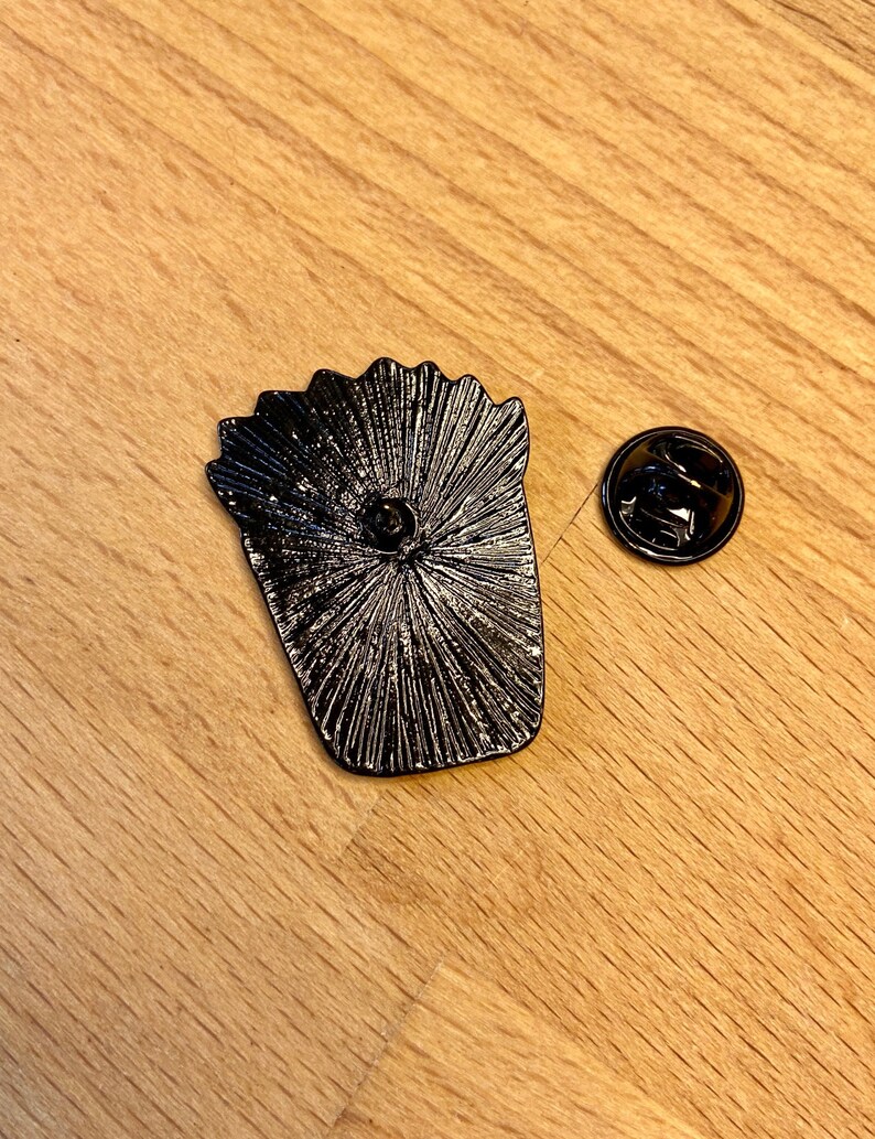 Puede incluir: Un pin de metal negro con forma de recipiente de patatas fritas, que muestra el reverso texturizado con un poste central y una pieza de cierre circular de metal negro separada, ambos con un acabado oscuro y mate.