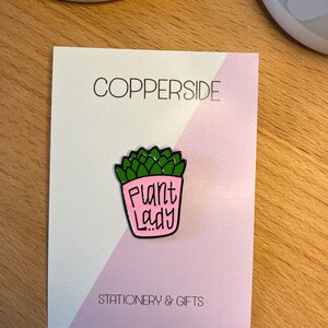 Puede incluir: Un pin de esmalte met&aacute;lico con forma de maceta rosa que contiene una planta suculenta verde. La maceta presenta el texto negro "Plant Lady" en una fuente manuscrita. El pin est&aacute; sujeto a una tarjeta de soporte blanca y rosa claro con la etiqueta "COPPERSIDE STATIONERY & GIFTS".