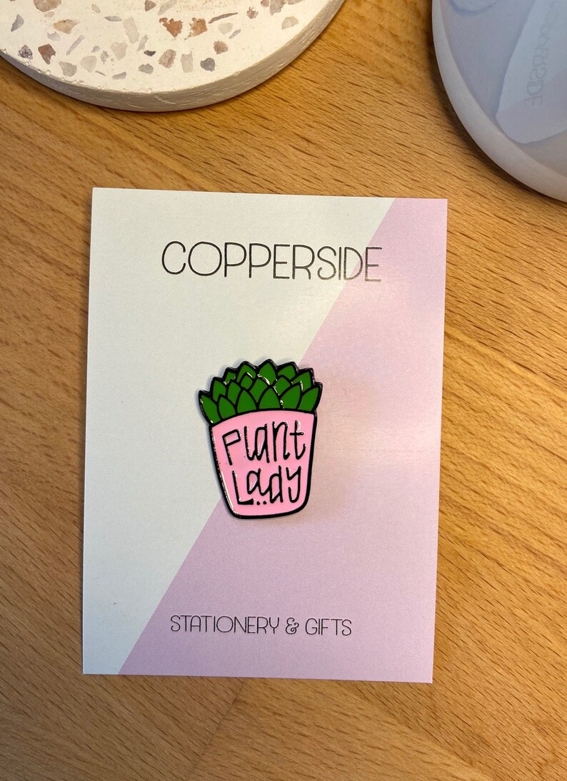 Puede incluir: Un pin de esmalte met&aacute;lico con forma de maceta rosa que contiene una planta suculenta verde. La maceta presenta el texto negro "Plant Lady" en una fuente manuscrita. El pin est&aacute; sujeto a una tarjeta de soporte blanca y rosa claro con la etiqueta "COPPERSIDE STATIONERY & GIFTS".