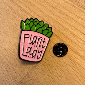 Puede incluir: Un pin de esmalte rosa con forma de suculenta en maceta con hojas verdes. La maceta rosa presenta el texto negro "Plant Lady" escrito en una fuente cursiva estilizada. Un cierre de metal negro separado se encuentra a la derecha del pin.
