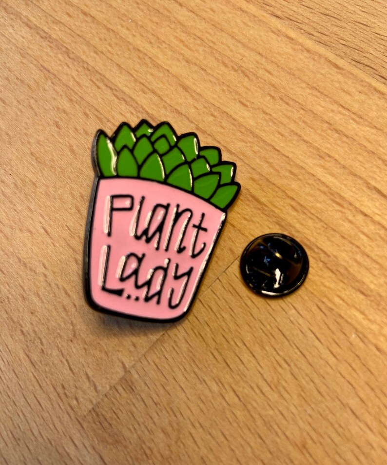 Puede incluir: Un pin de esmalte rosa con forma de suculenta en maceta con hojas verdes. La maceta rosa presenta el texto negro "Plant Lady" escrito en una fuente cursiva estilizada. Un cierre de metal negro separado se encuentra a la derecha del pin.