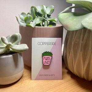 Puede incluir: Un pin de esmalte met&aacute;lico con forma de suculenta en maceta rosa con hojas verdes y el texto "PLANT LADY" en letras negras. El pin est&aacute; sujeto a una tarjeta de presentaci&oacute;n rectangular con el texto "COPPERSIDE" y "STATIONERY & GIFTS".