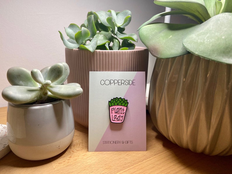 Puede incluir: Un pin de esmalte met&aacute;lico con forma de suculenta en maceta rosa con hojas verdes y el texto "PLANT LADY" en letras negras. El pin est&aacute; sujeto a una tarjeta de presentaci&oacute;n rectangular con el texto "COPPERSIDE" y "STATIONERY & GIFTS".