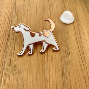 Puede incluir: Un broche de metal esmaltado en color oro rosa con forma de perro de pie, con relleno de esmalte blanco y dos manchas metálicas de color oro rosa en el lomo. El perro tiene la cola curvada y un contorno sencillo. Un cierre de goma blanco está al lado.