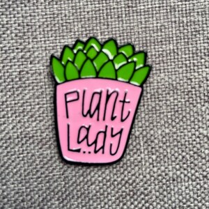 Puede incluir: Un pin de esmalte met&aacute;lico que muestra una planta suculenta de color verde brillante en una maceta rosa claro. La maceta tiene el texto Plant Lady escrito en letras negras estilizadas a mano. El pin tiene un borde de metal negro alrededor.