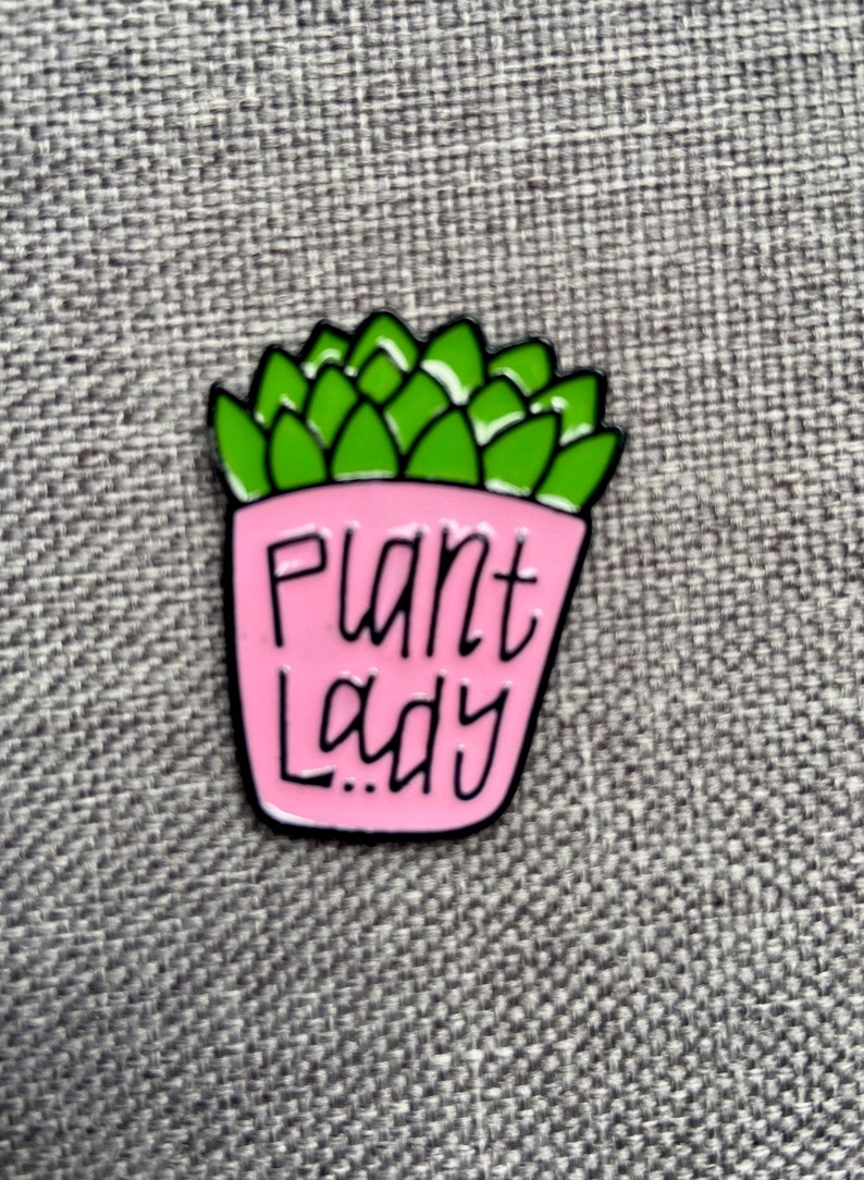 Puede incluir: Un pin de esmalte met&aacute;lico que muestra una planta suculenta de color verde brillante en una maceta rosa claro. La maceta tiene el texto Plant Lady escrito en letras negras estilizadas a mano. El pin tiene un borde de metal negro alrededor.