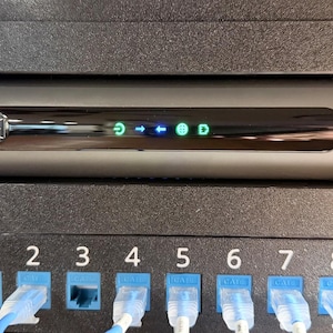 May include: A silver Motorola cable modem with a black front panel featuring five illuminated status icons, mounted in a black rack above a network patch panel with ten numbered ports, several of which are connected with white and blue ethernet cables.