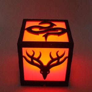 May include: A wooden cube-shaped lamp with a red light inside. The lamp has a black silhouette of a deer head with antlers on the front and a snake design on the top.