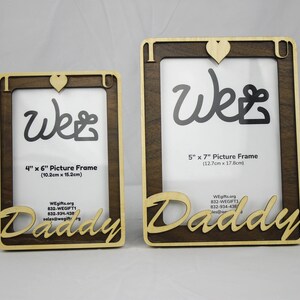 May include: Two wooden picture frames with a heart and the word "We" in cursive. The larger frame is 5 inches by 7 inches (12.7 cm by 17.8 cm) and the smaller frame is 4 inches by 6 inches (10.2 cm by 15.2 cm). Both frames have the word "Daddy" in cursive below the picture opening.