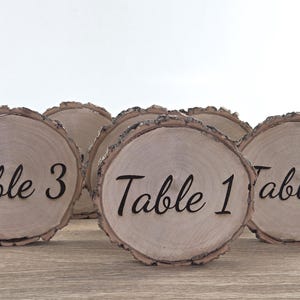 Puede incluir: Conjunto de portatarjetas de mesa rústicos de madera. Cada rodaja de madera circular tiene un borde de corteza natural y presenta la palabra "Mesa" seguida de un número en fuente negra. Ideal para bodas o eventos.