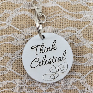 May include: White round keychain with a silver key ring. The keychain has black text that reads "Think Celestial" with a decorative swirl design.