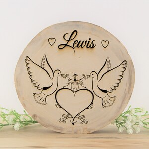 Puede incluir: Un letrero redondo de madera con el nombre "Lewis" grabado en letra cursiva. Dos palomas se miran con un corazón en el medio. Las palomas sostienen una cinta con un corazón en ella. El letrero está decorado con corazones y espirales.